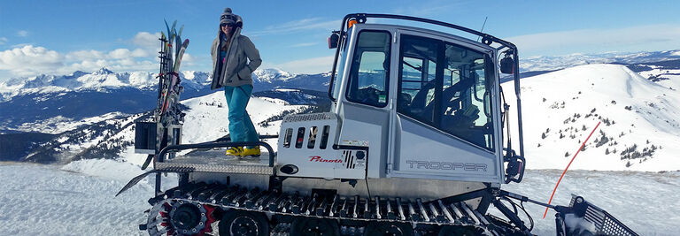 Erika standing on the back of a snow cat next to skis in front of a mountain landscape
